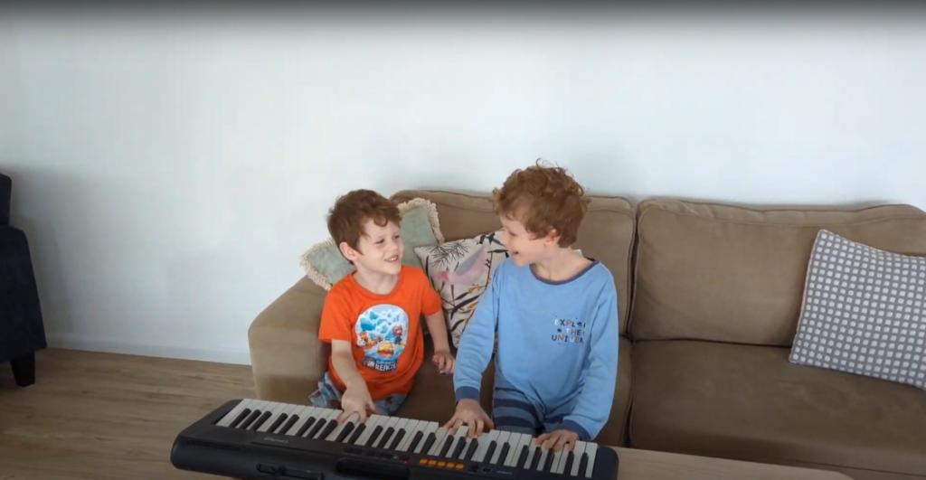 Two small boys giggle at each other while playing on a piano keyboard together.