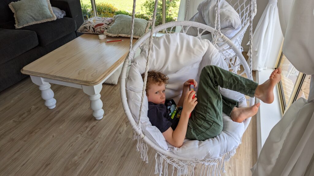 A boy is curled up in a swing chair, while holding a Tablet.